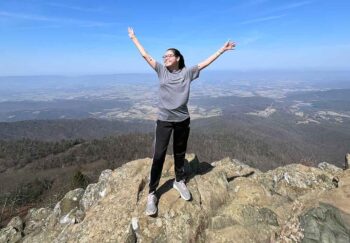A young transplant survivor celebrates with arms out on top of a mountain.
